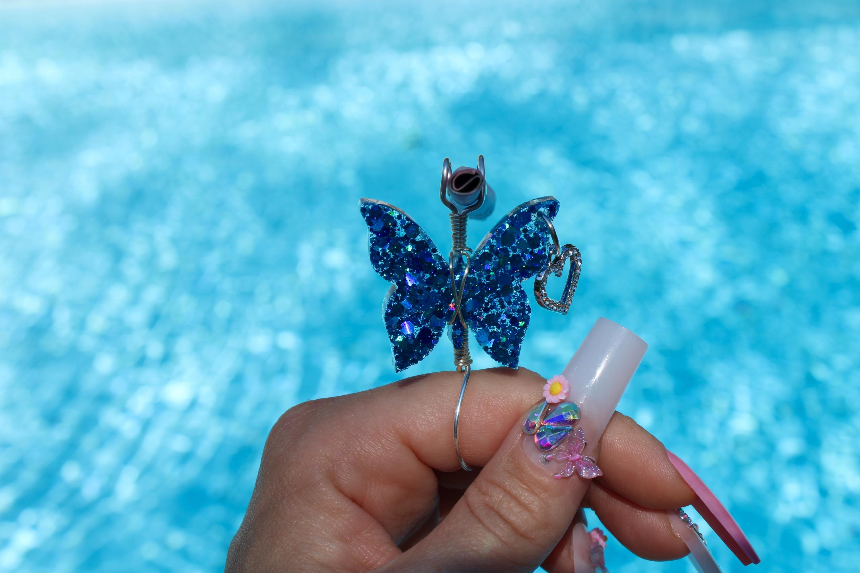 Woman holds a glittery blue butterfly roach clip above a pool, showcasing long, decorated acrylic nails.