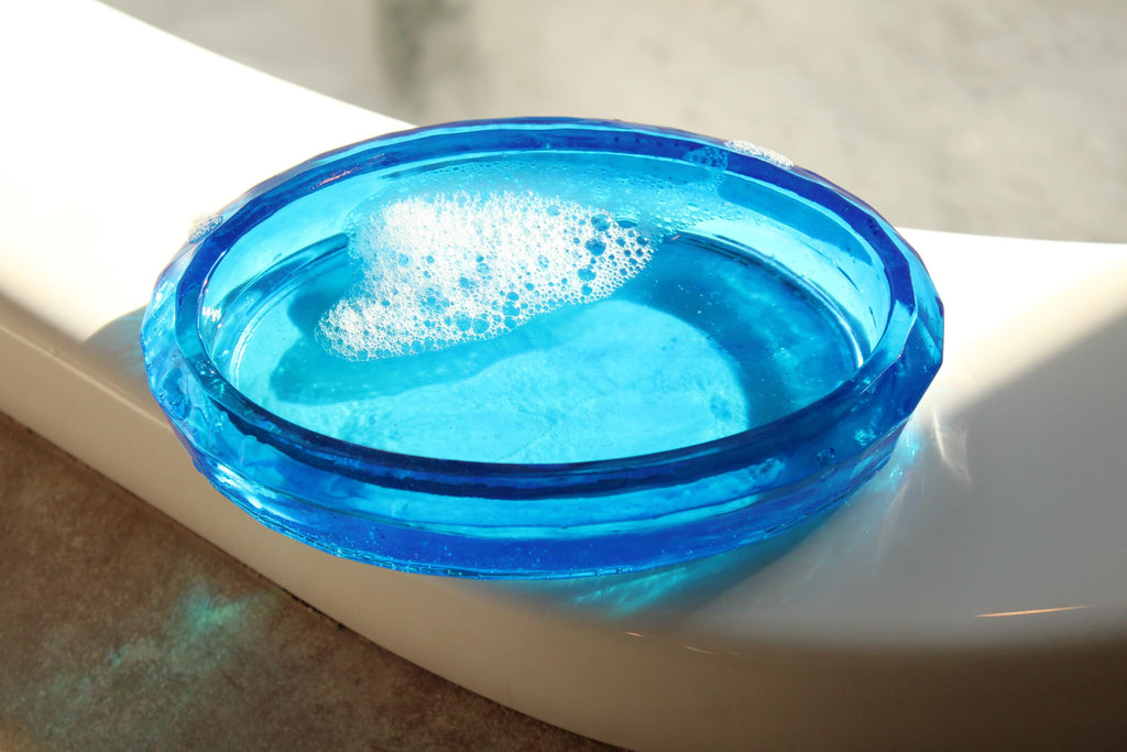 Sunlight illuminates a decorative, blue soap dish filled with water and bubbles on a white bathroom surface.
