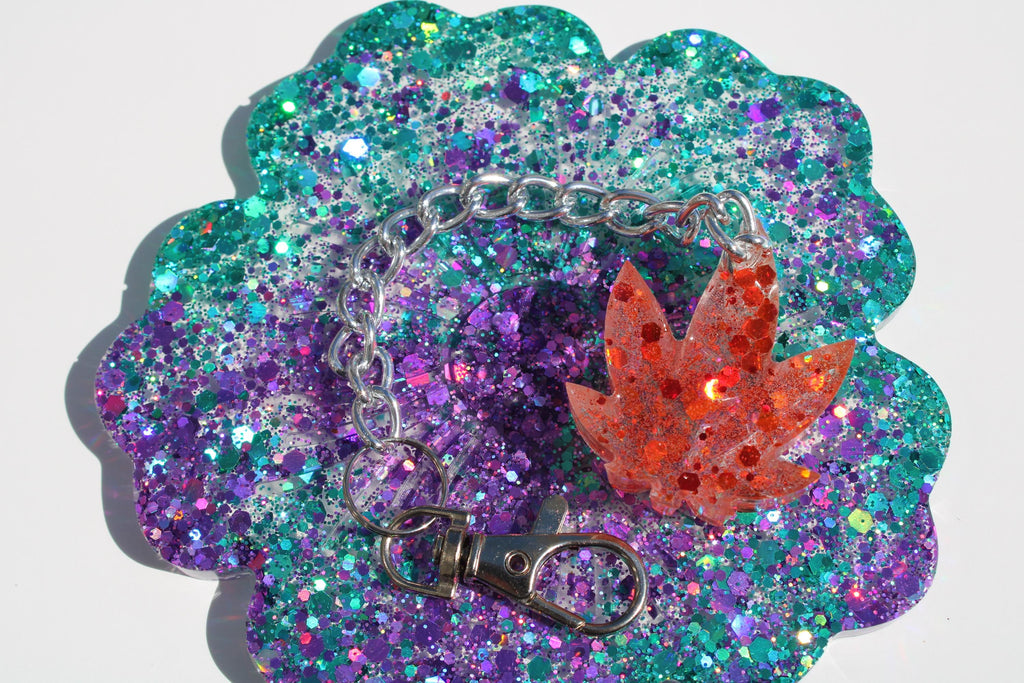 A sparkly handmade resin keychain featuring a red glitter weed leaf charm sits on a glittery turquoise and purple coaster.