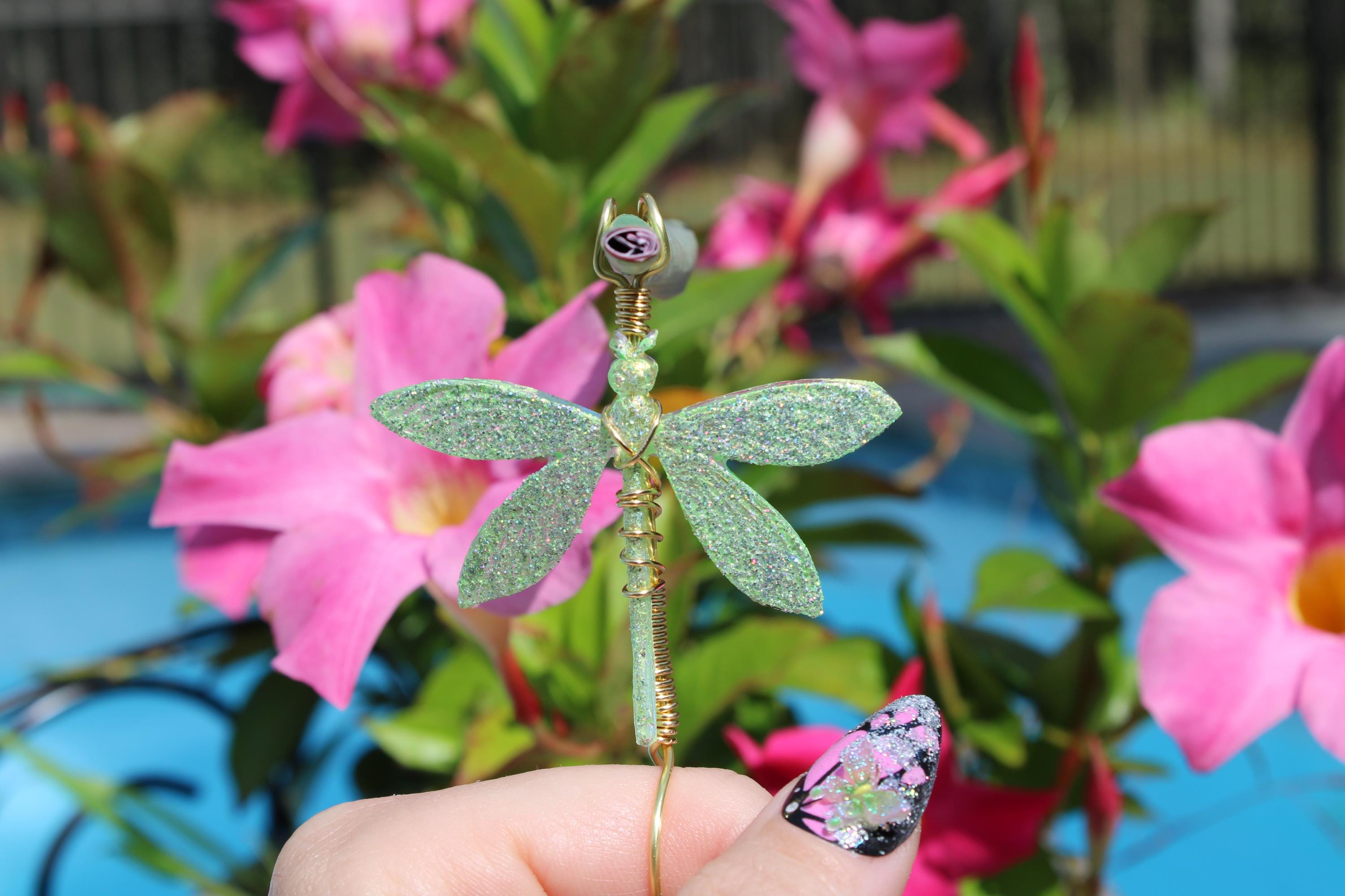 A hand with decorative nail art holds a sparkly green dragonfly smoke accessory in front of pink mandevilla flowers.