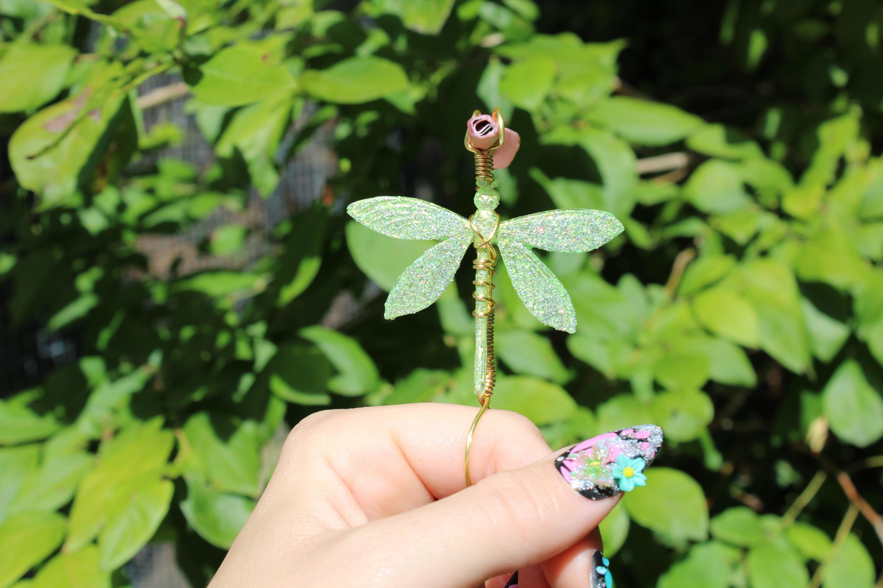 A hand with floral nail art holds a sparkly green dragonfly smoke ring with a pink rose accent against a leafy green background.