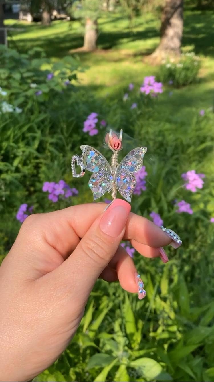 A hand with pink nails holds a sparkly butterfly smoke ring featuring a heart charm, posing it against a garden backdrop of purple flowers.