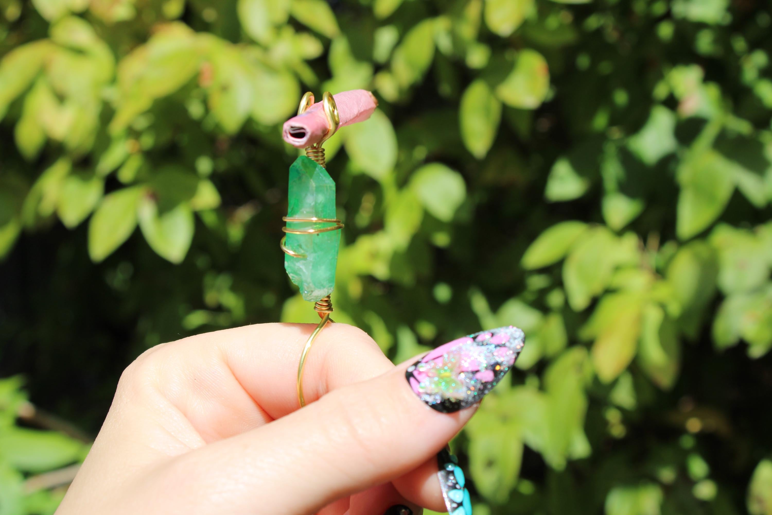 A hand with artistically painted nails holds a handcrafted crystal smoke ring featuring a green quartz crystal.