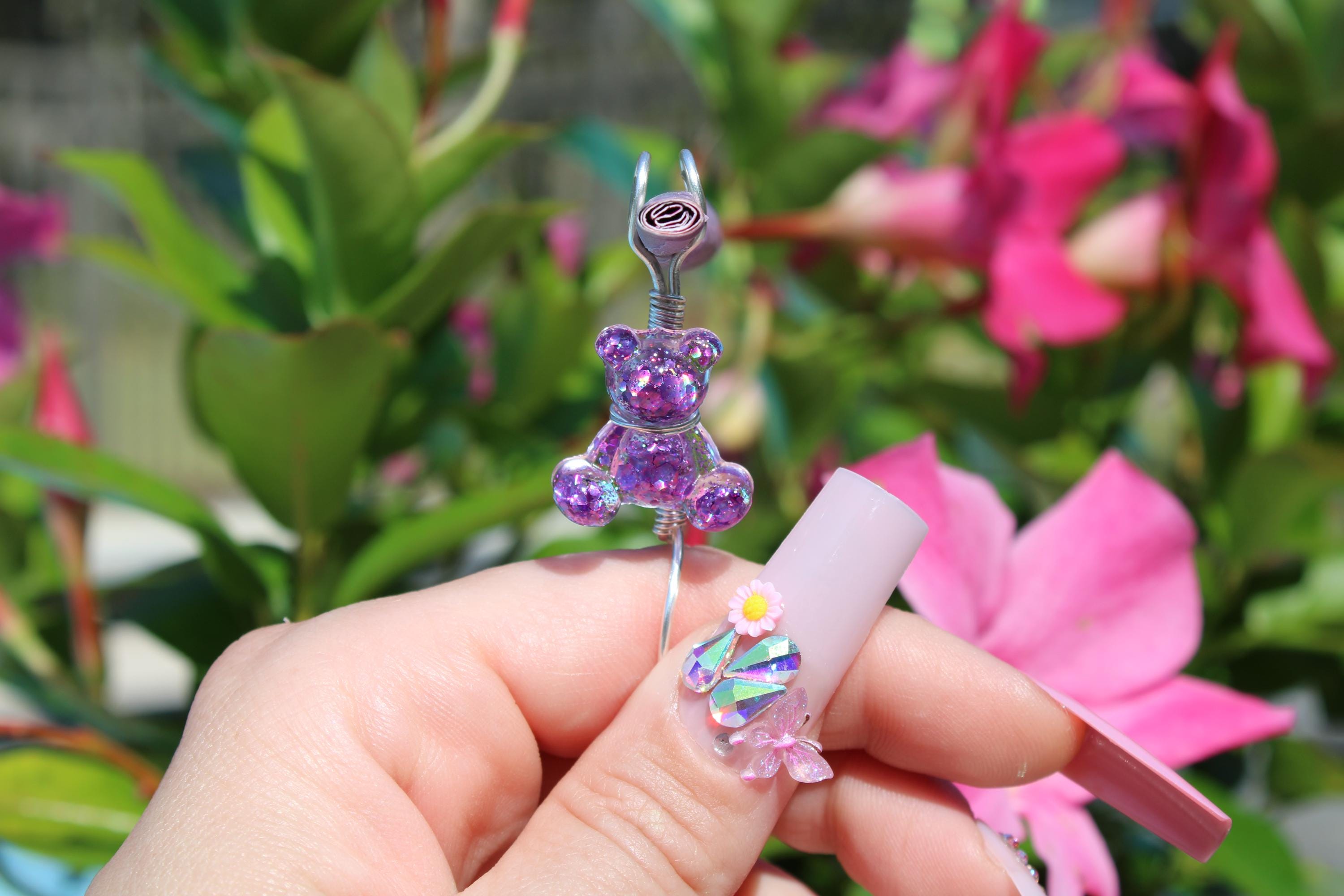 A hand displays an elaborate nail art design, featuring a purple teddy bear smoke ring, flower embellishments, and iridescent crystals against a background of pink blooms.