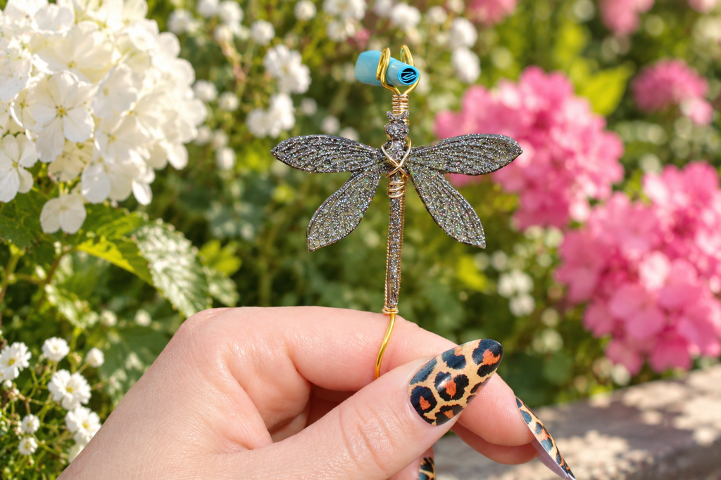 A hand with leopard-print nail art holds a wire-wrapped dragonfly smoke ring against a blurred garden background of white and pink flowers.