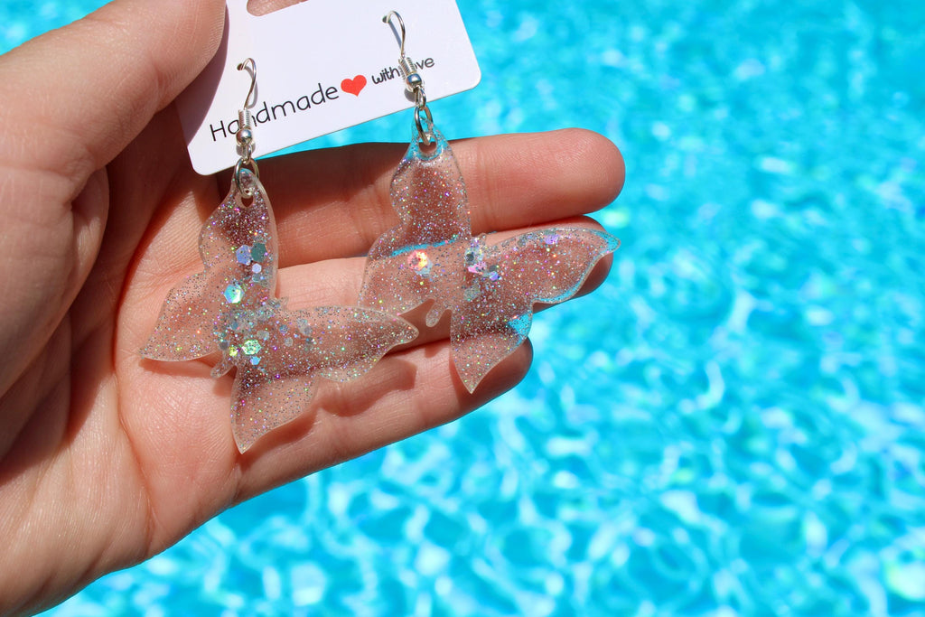A hand displays sparkly, handmade butterfly earrings against a blurred pool background.