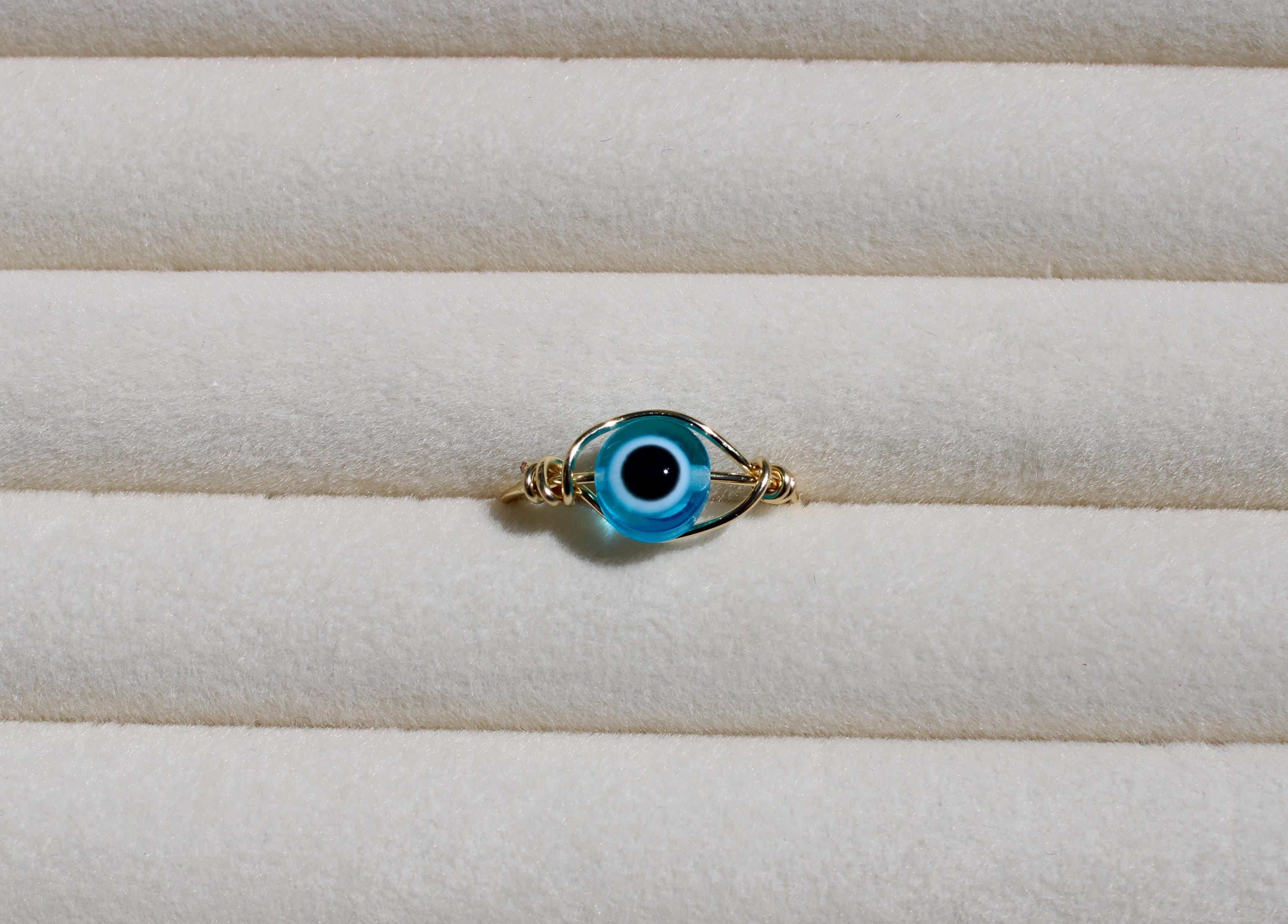 A handmade, wire-wrapped ring showcases a light blue evil eye bead on a textured, light-colored surface.