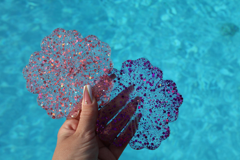 Hand holding two glittery, flower-shaped drink coasters against a blue water background