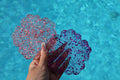Hand holding two glittery, flower-shaped drink coasters against a blue water background