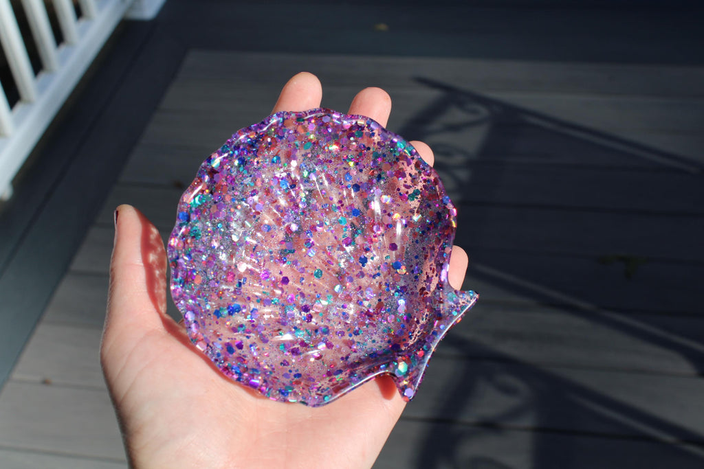 Hand holding a glittery purple and pink seashell soap dish on a wooden deck
