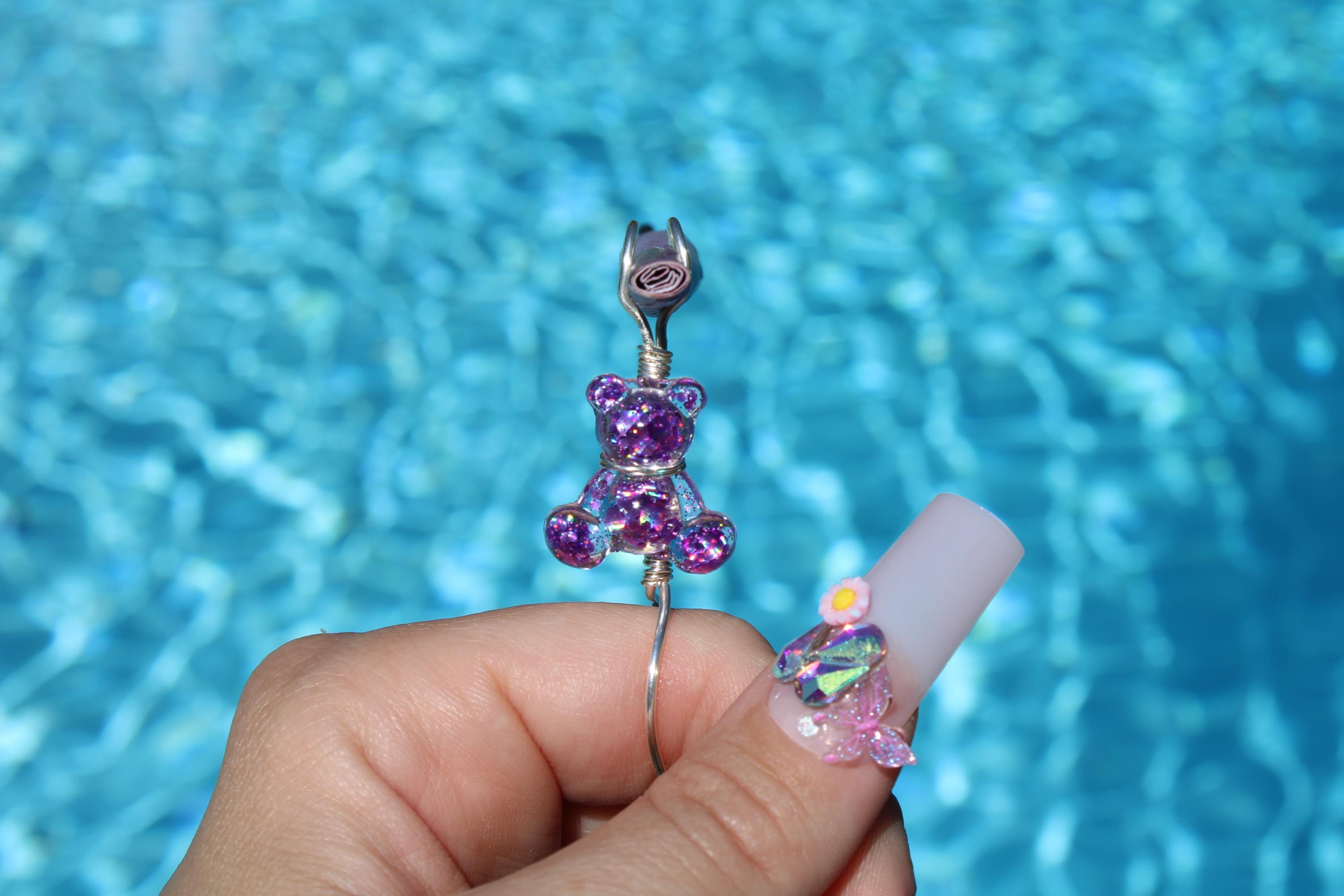 A hand displays a whimsical, sparkly purple teddy bear smoke ring and a creatively decorated, long acrylic nail against a blurred blue pool background.
