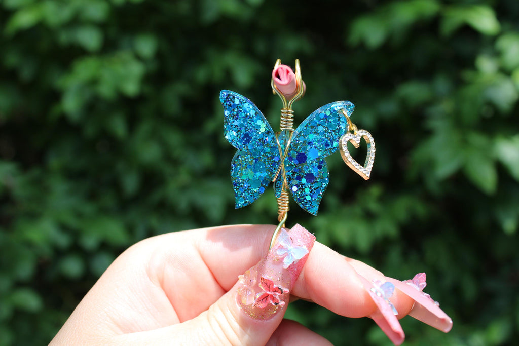 A hand with elaborate butterfly-themed nail art presents a sparkly blue butterfly smoke ring featuring a rhinestone heart charm.