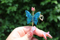 A hand with elaborate butterfly-themed nail art presents a sparkly blue butterfly smoke ring featuring a rhinestone heart charm.
