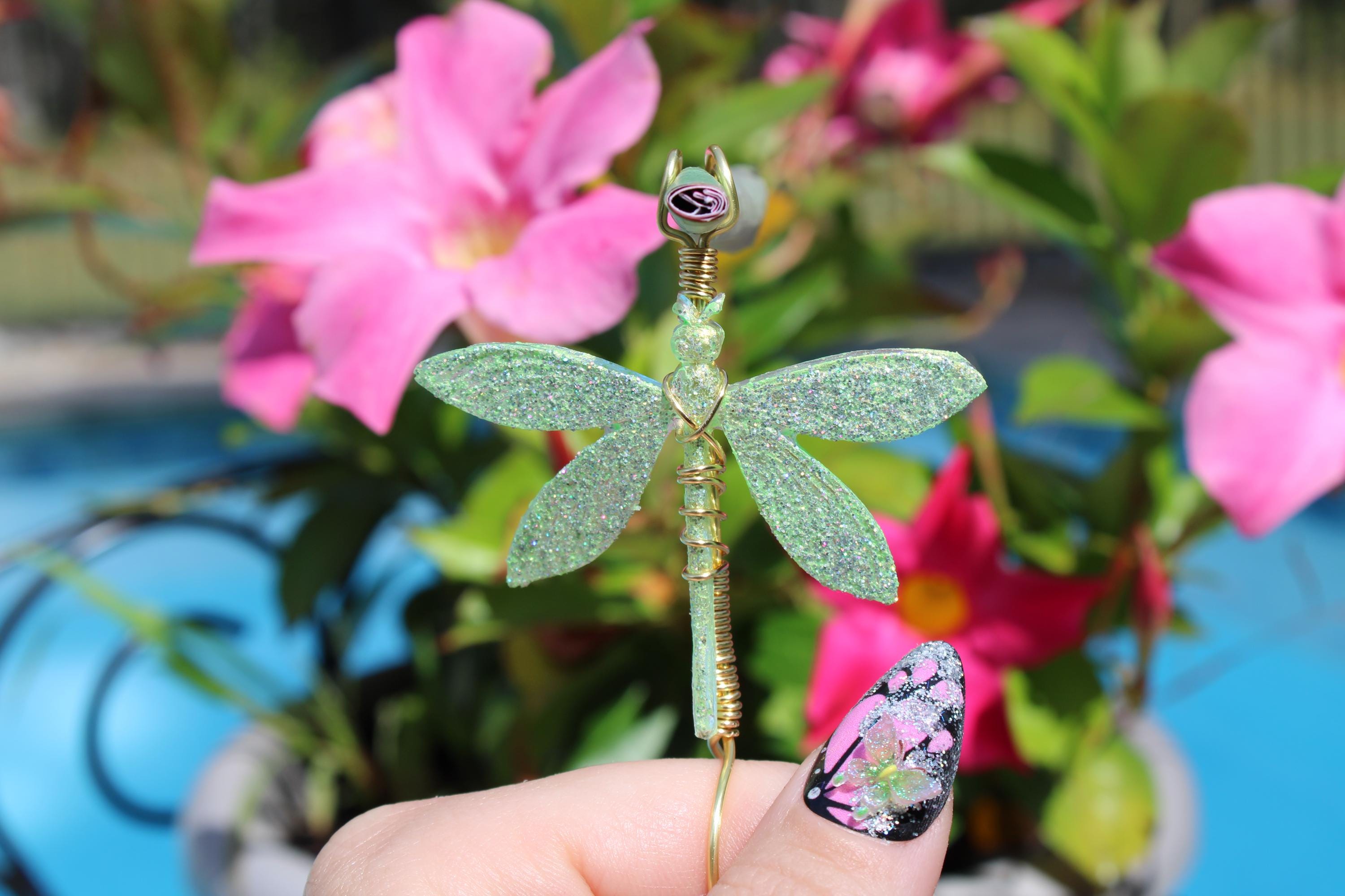 A hand with a decorative nail holds a sparkly green and gold dragonfly smoke ring in front of pink flowers.