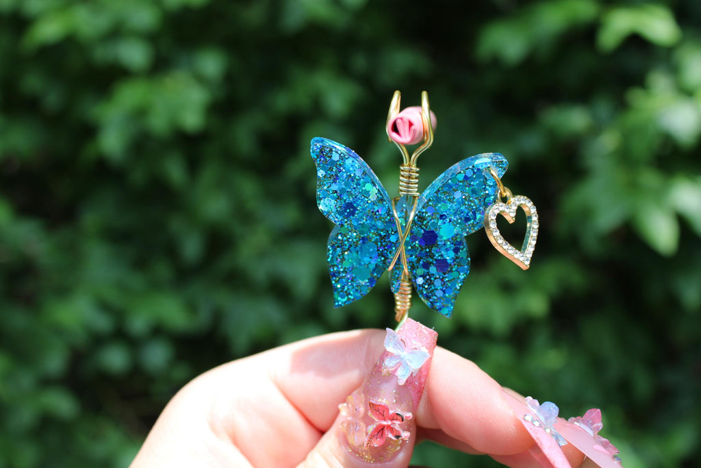 A hand with elaborate nail art holds a sparkly blue resin butterfly smoke ring with a rhinestone heart charm.