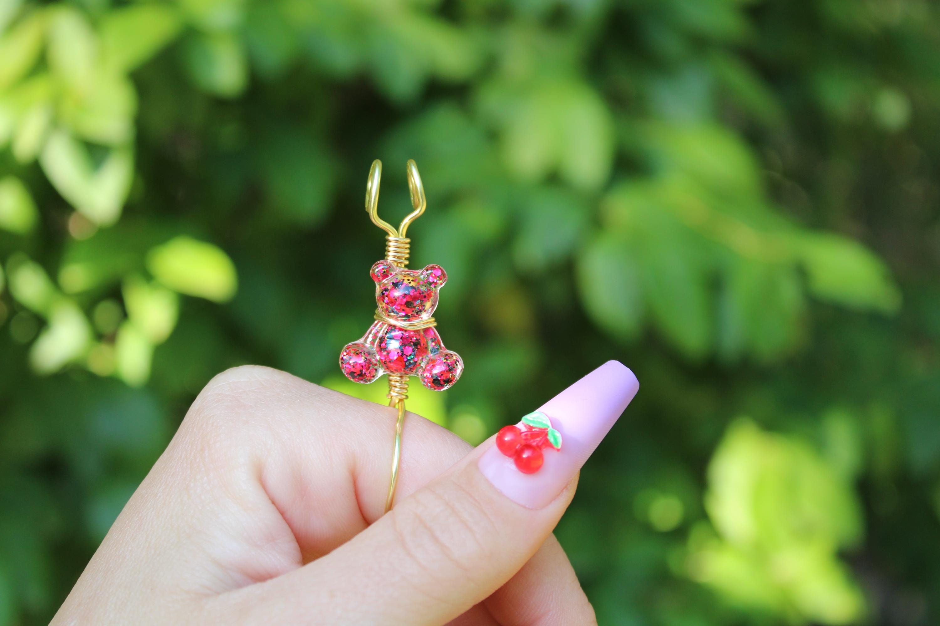 A hand with a cherry-decorated nail holds a pink sequin teddy bear smoke ring.