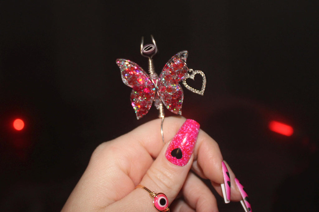 Butterfly-shaped smoke ring with red glittery wings held by a hand with pink nail polish against a dark background.