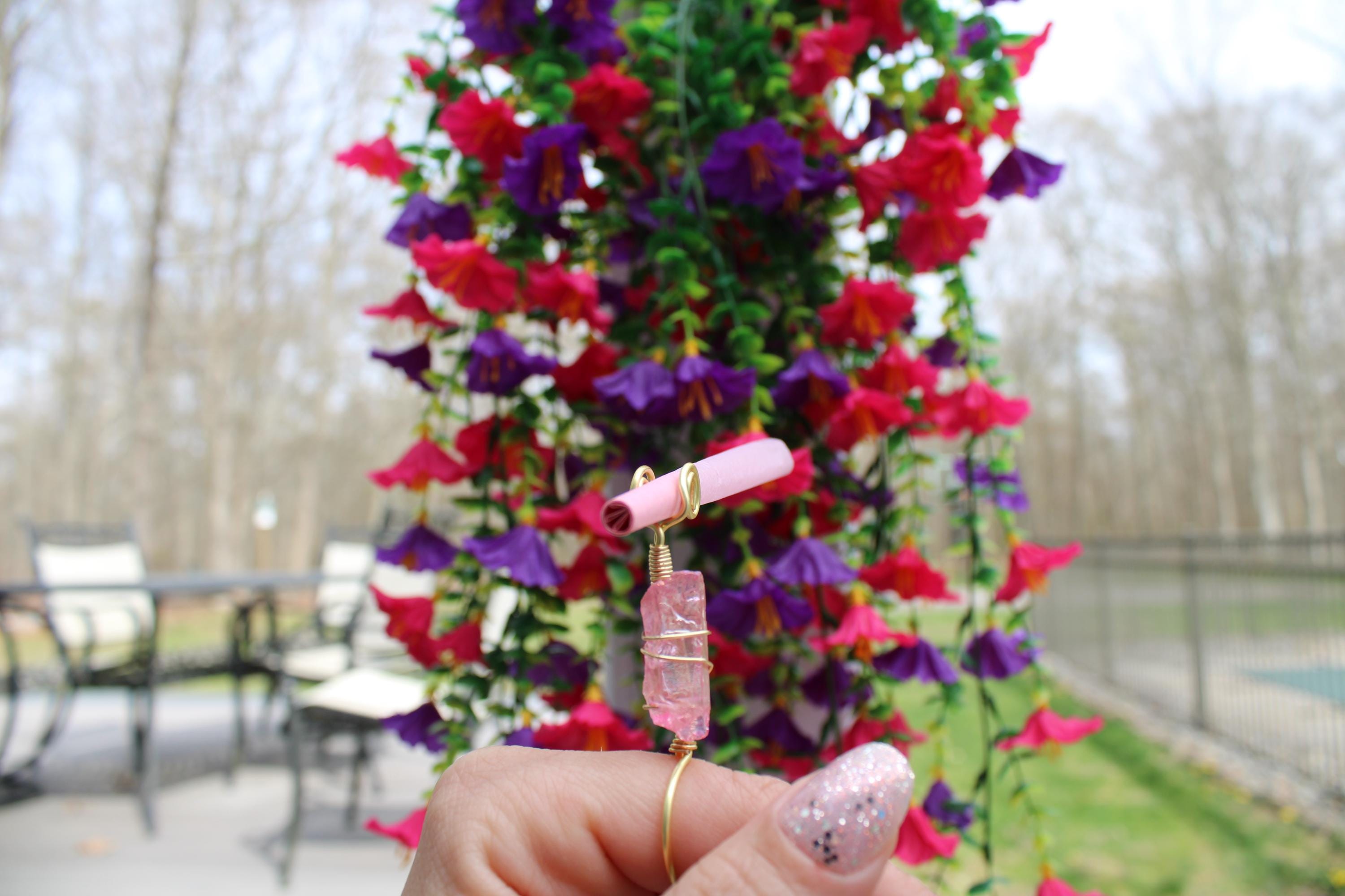 A hand models a gold colored wire wrapped rose quartz crystal smoke ring.