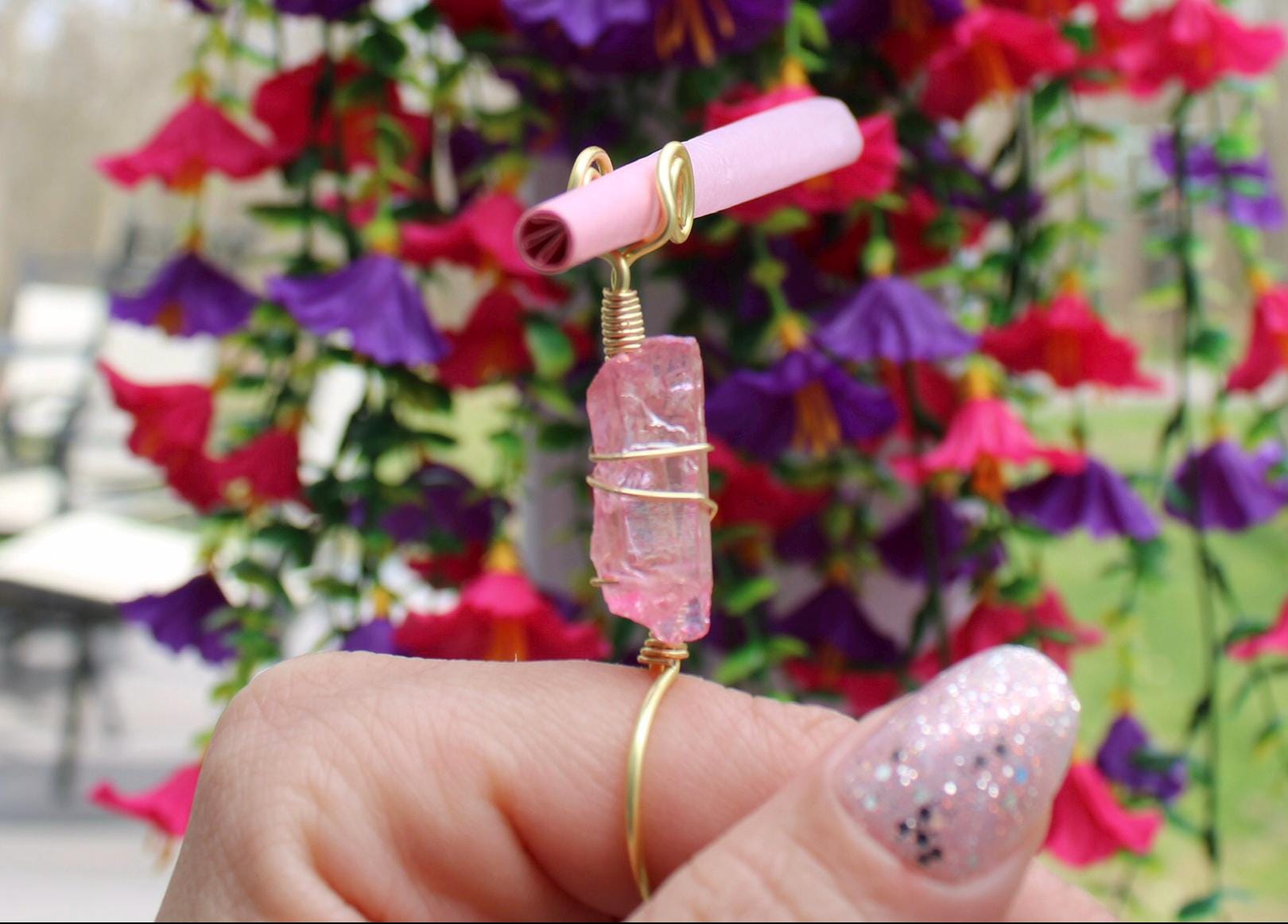 A hand with glittery pink nails displays a handmade crystal smoke ring featuring a rough-cut pink gemstone and a small pink rolled paper accessory.