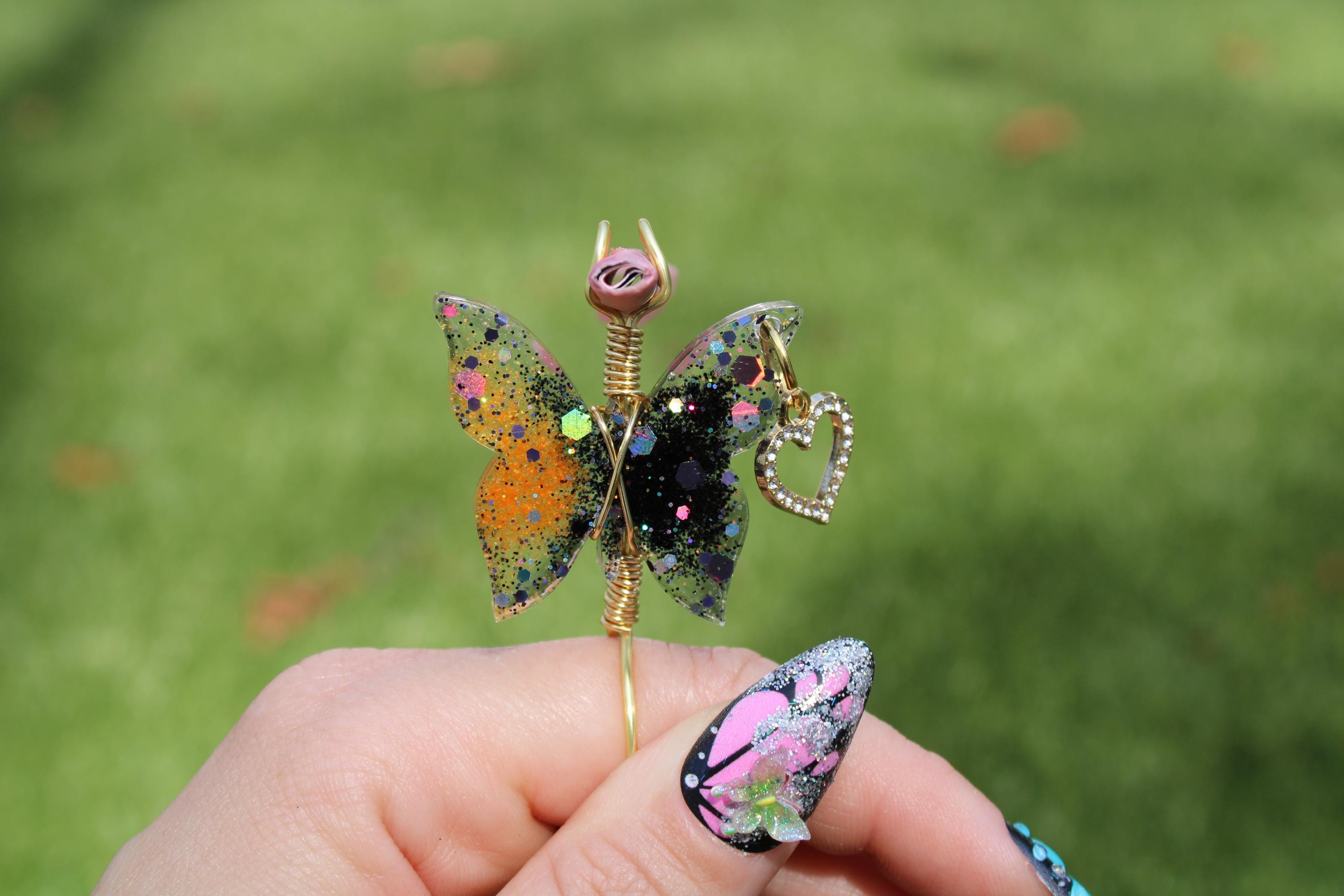 A hand with a butterfly-themed manicure holds up a sparkly butterfly smoke ring against a green background.