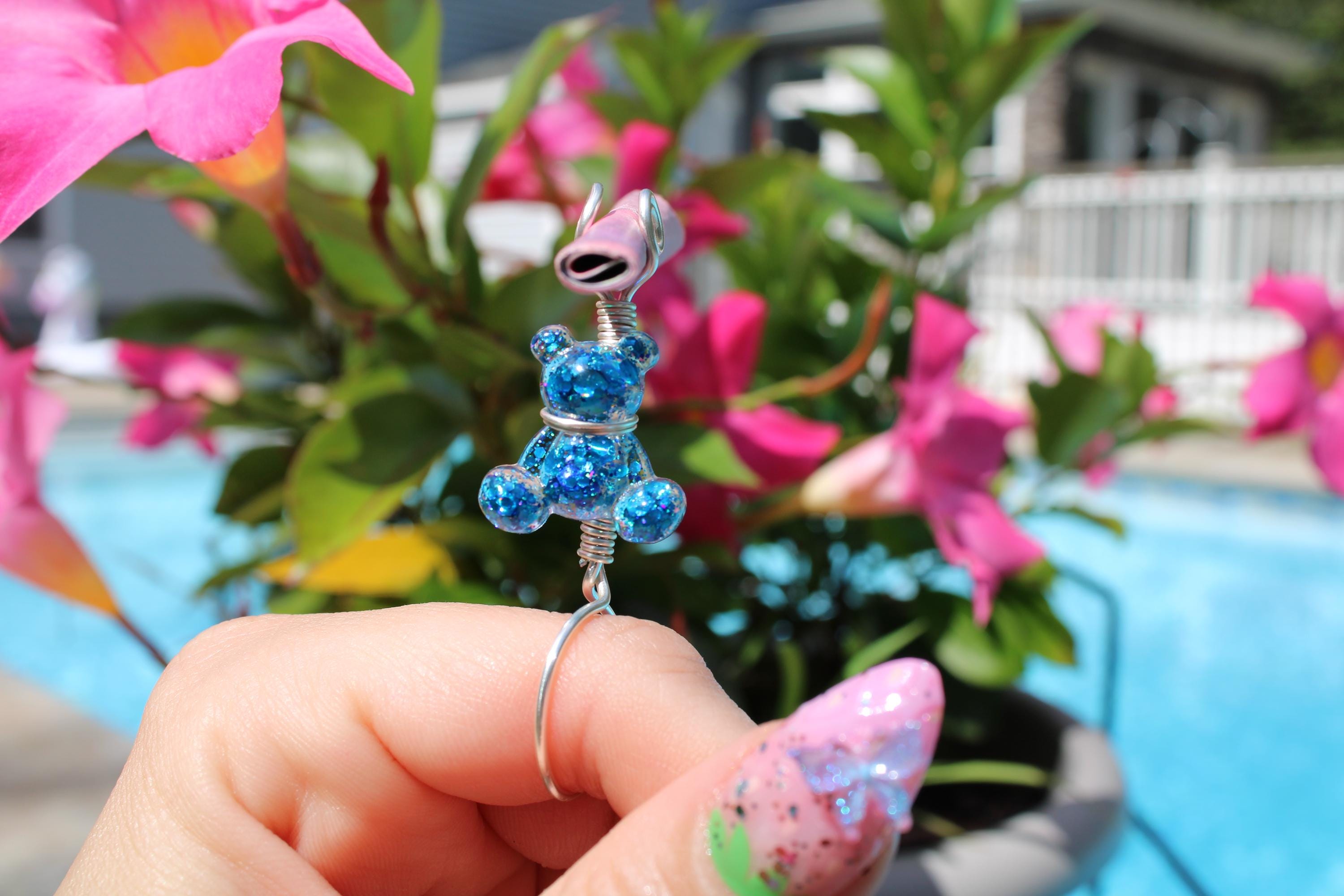A manicured hand holds a blue sequin teddy bear smoke ring against a flowery background by a swimming pool.