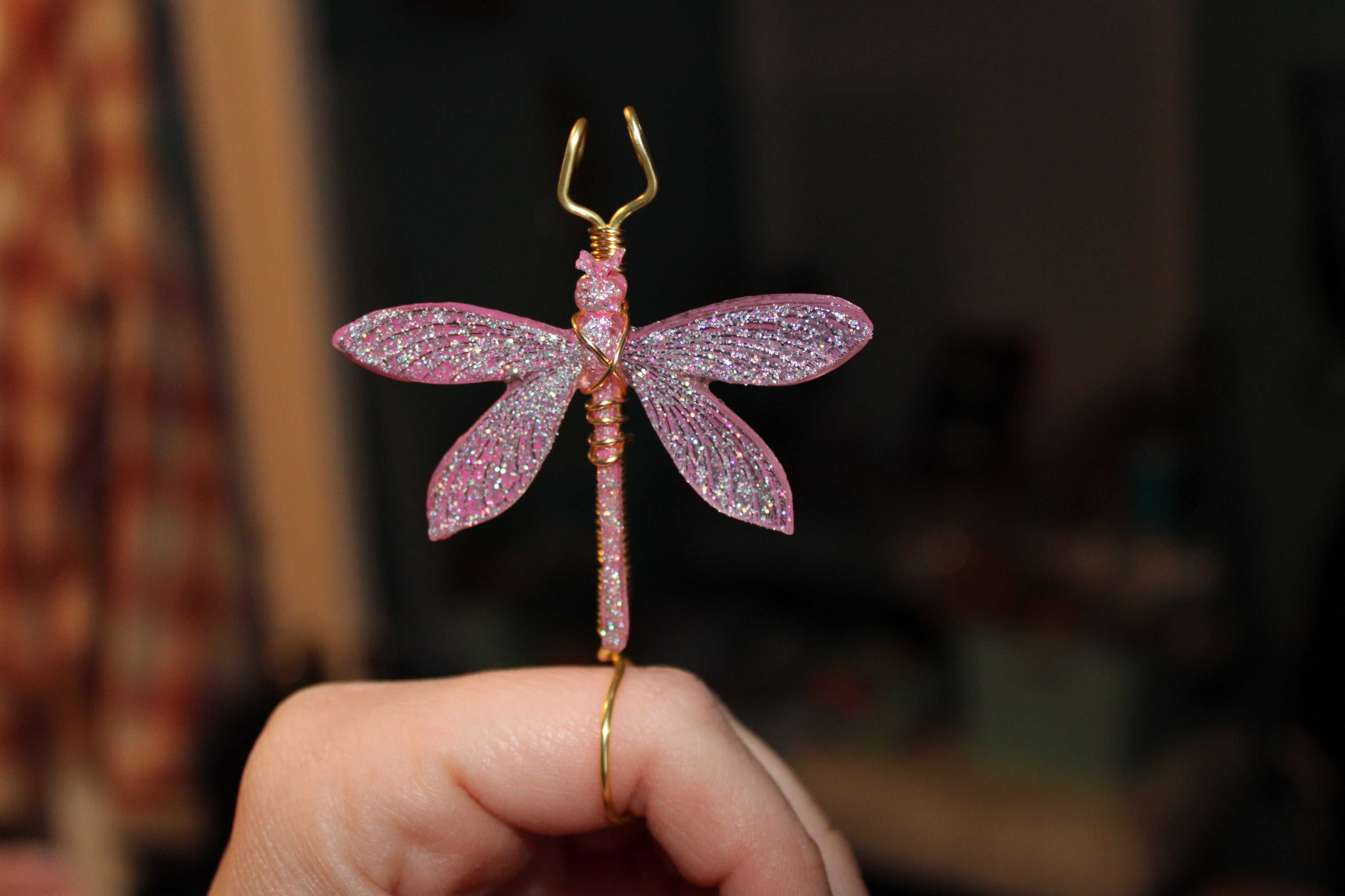 A hand holds a sparkly pink and silver dragonfly smoke ring with gold wire.
