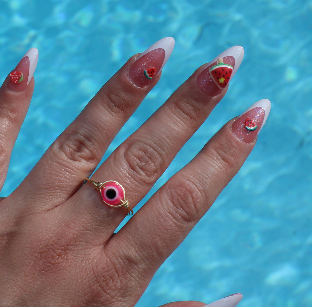 A woman's hand, sporting watermelon-themed nail art and a pink evil eye ring, is displayed against a blue pool backdrop.