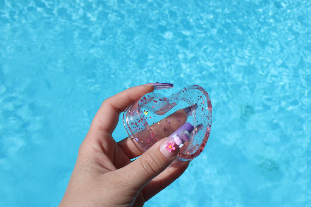 Hand holding a clear ring dish with pink glittery accents against a blue water background