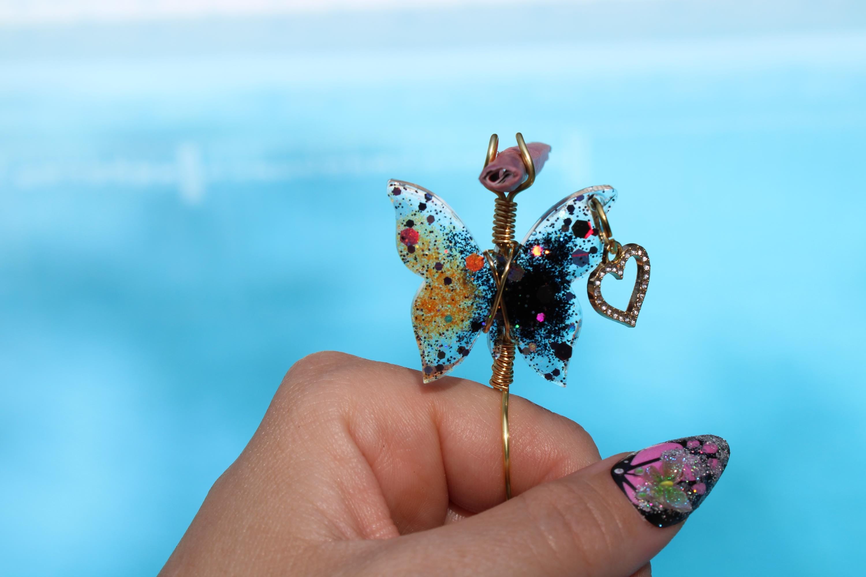 A hand displays a colorful, glittery resin butterfly smoke ring with a heart charm against a blue background.