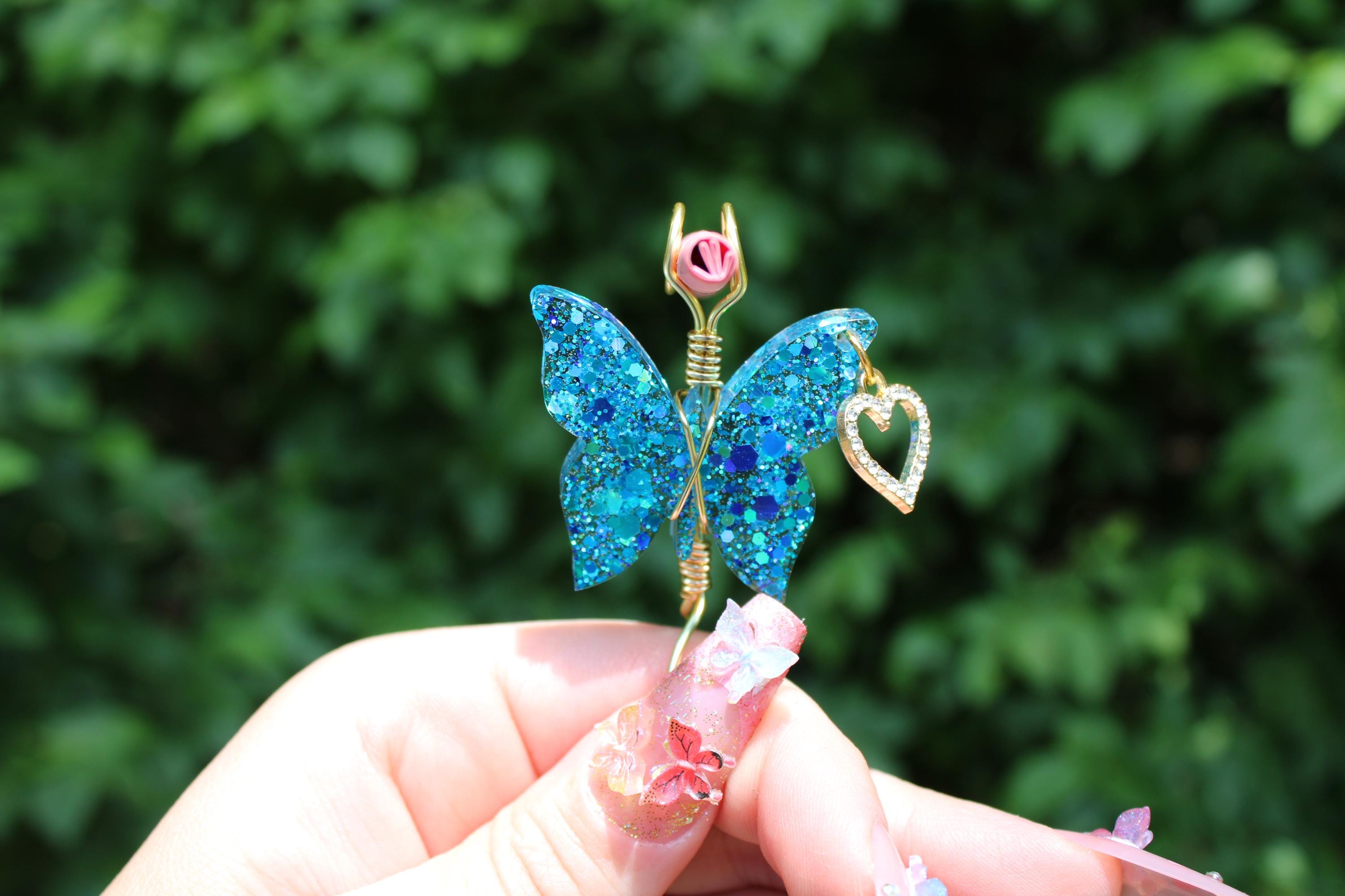 Hand showcases a shimmering blue glitter butterfly smoke ring featuring a gold heart charm.