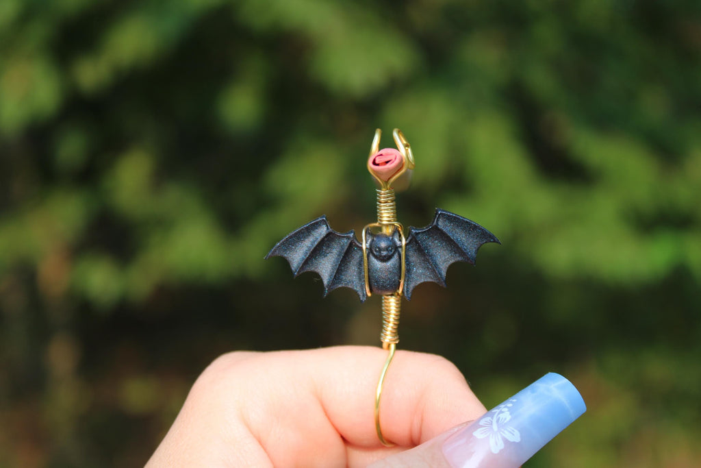 A hand displays a unique handmade gold wire smoke ring featuring a black bat  against a green background.