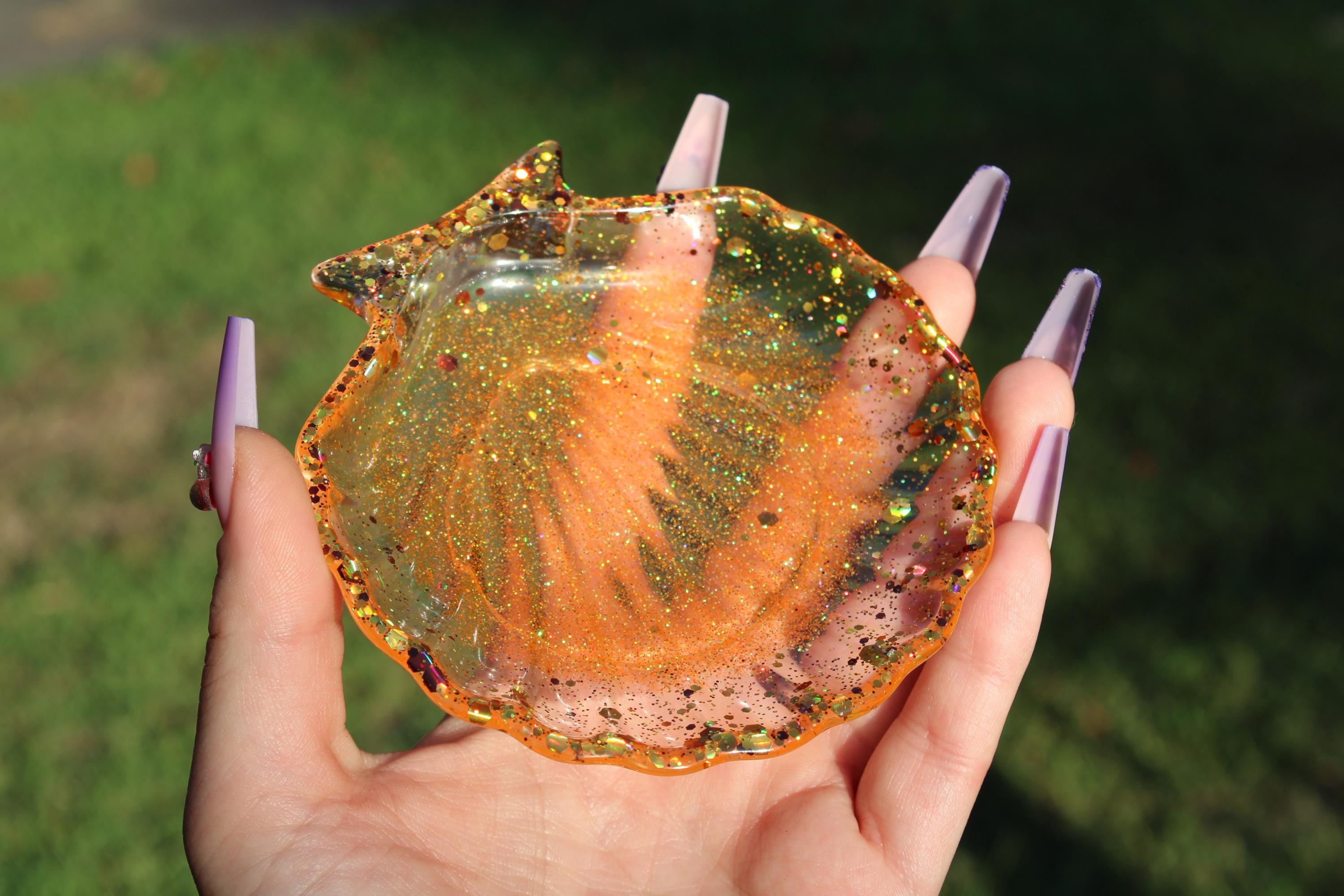 A hand with long, purple nails displays a sparkly, orange, seashell-shaped resin trinket dish.