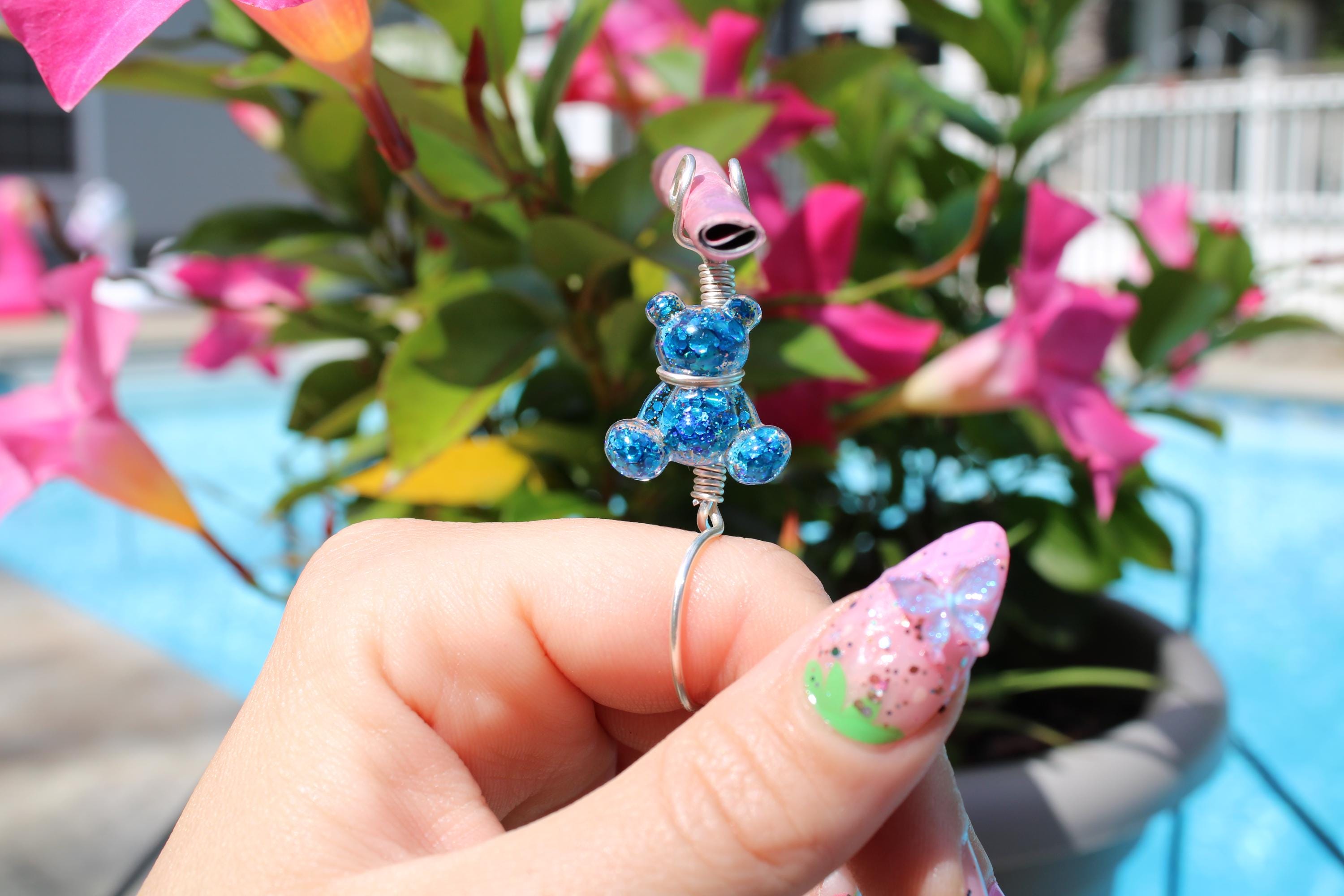 Woman's manicured hand holds a blue glitter teddy bear smoke ring against a background of pink mandevilla flowers and a blue pool.