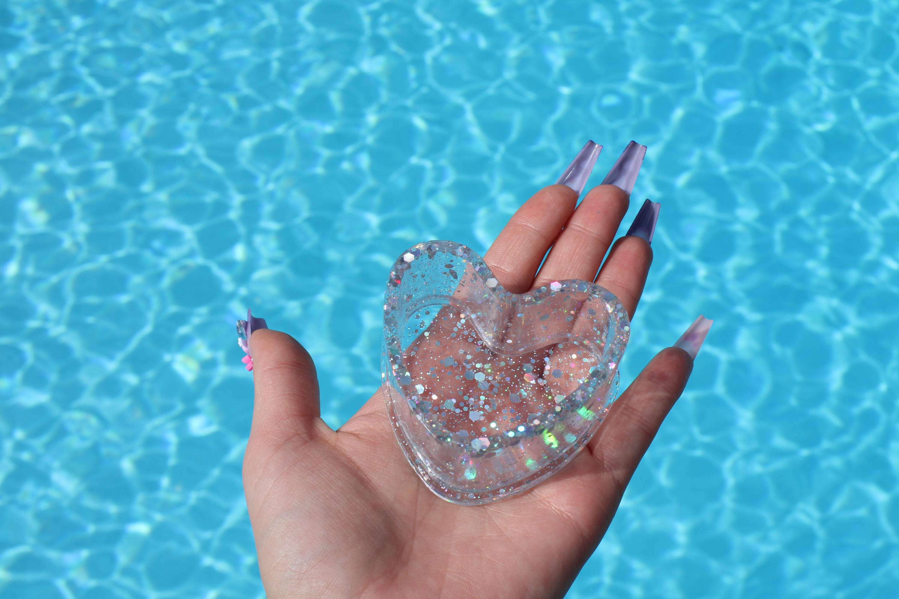 Heart-shaped glittery ring dish held by a hand with purple nail polish against a blue water background