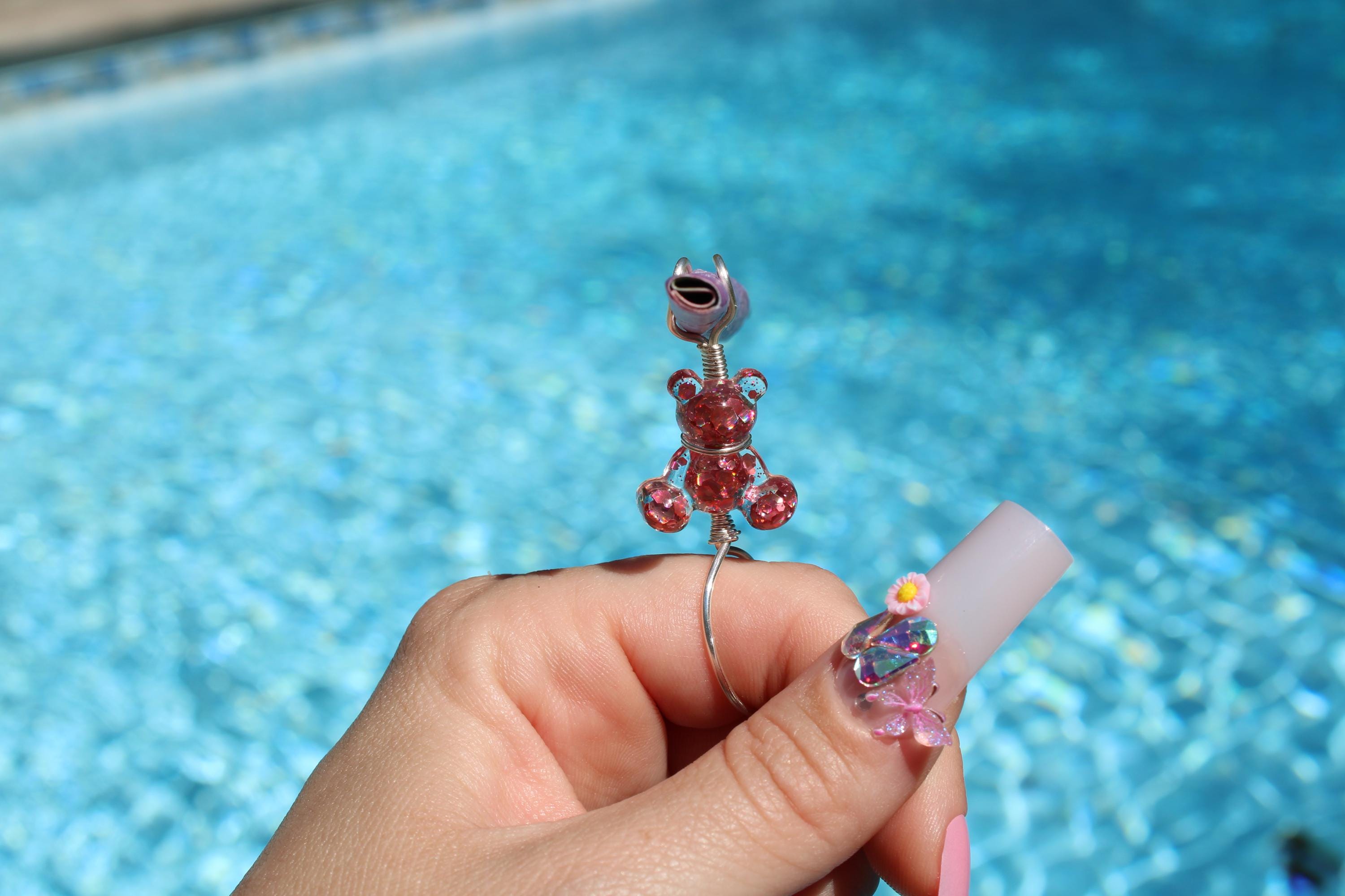 A hand showcases a unique handmade wire smoke ring featuring a pink sequin teddy bear with a swimming pool in the background.