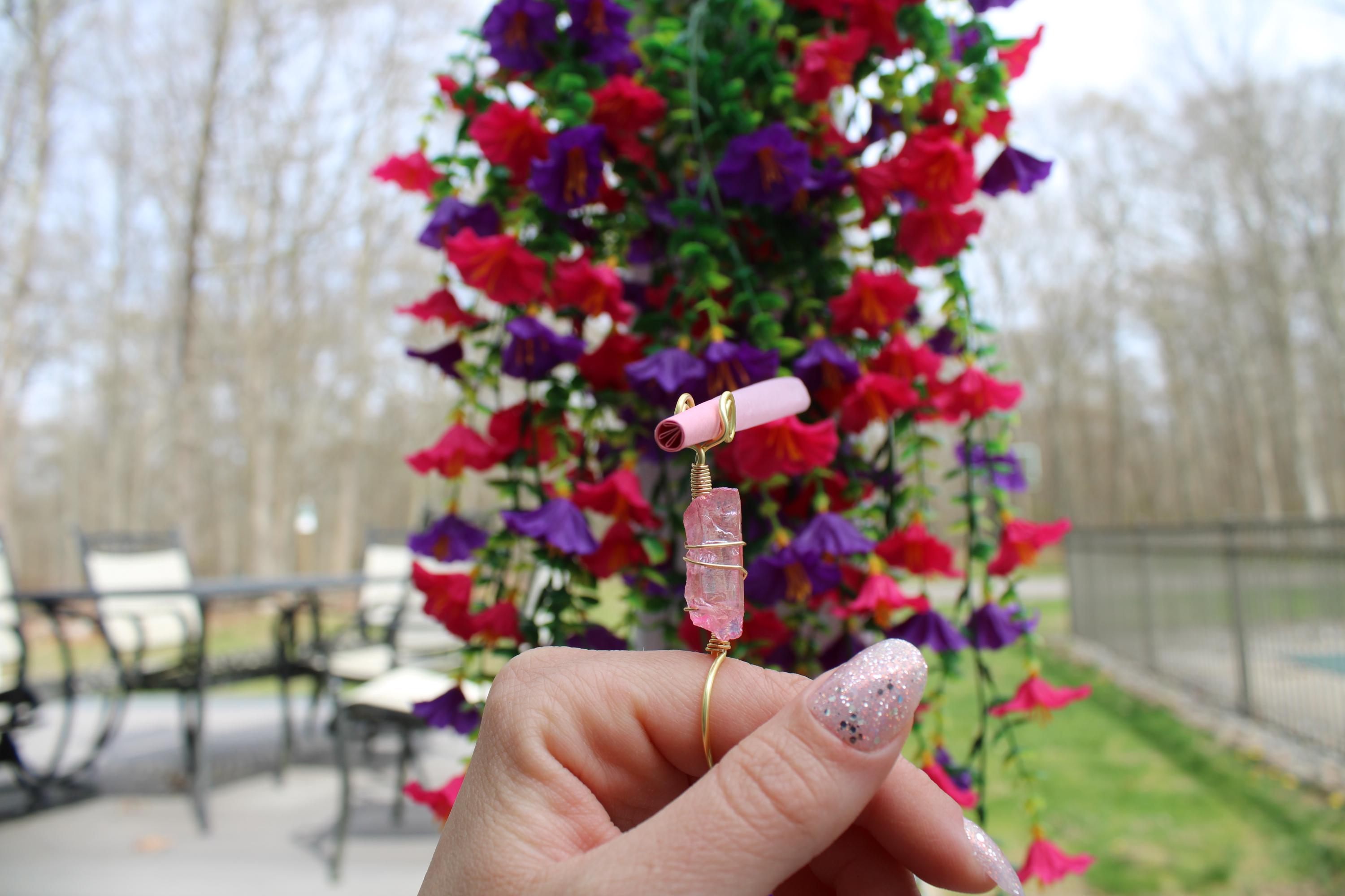 A hand displays a handmade rose quartz crystal smoke ring in front of flowers.