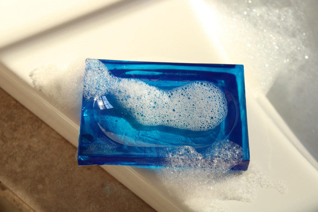 A blue resin soap dish holds white soap bubbles in a brightly lit bathroom.