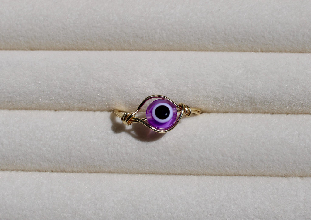 A handcrafted wire ring features a purple evil eye bead against a soft, cream-colored surface.