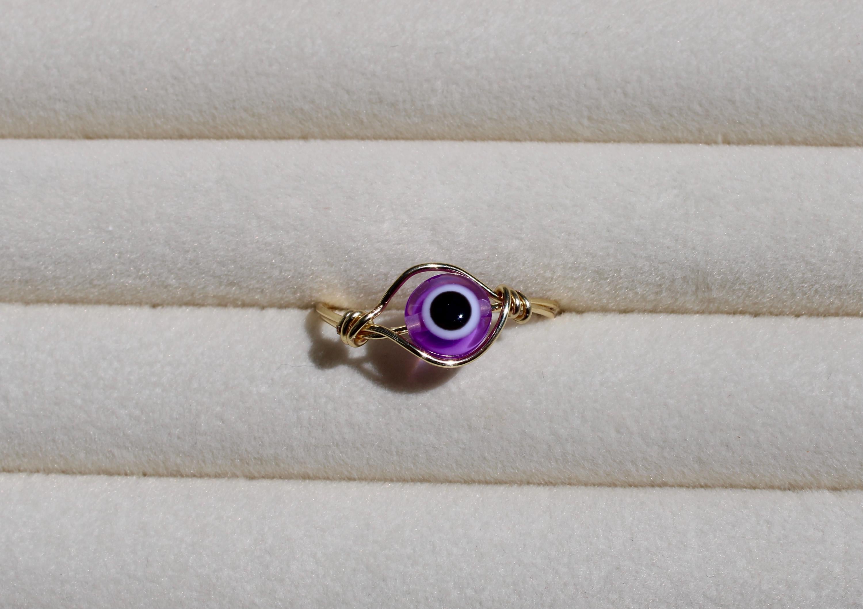 A handcrafted wire ring features a purple evil eye bead against a soft, cream-colored surface.