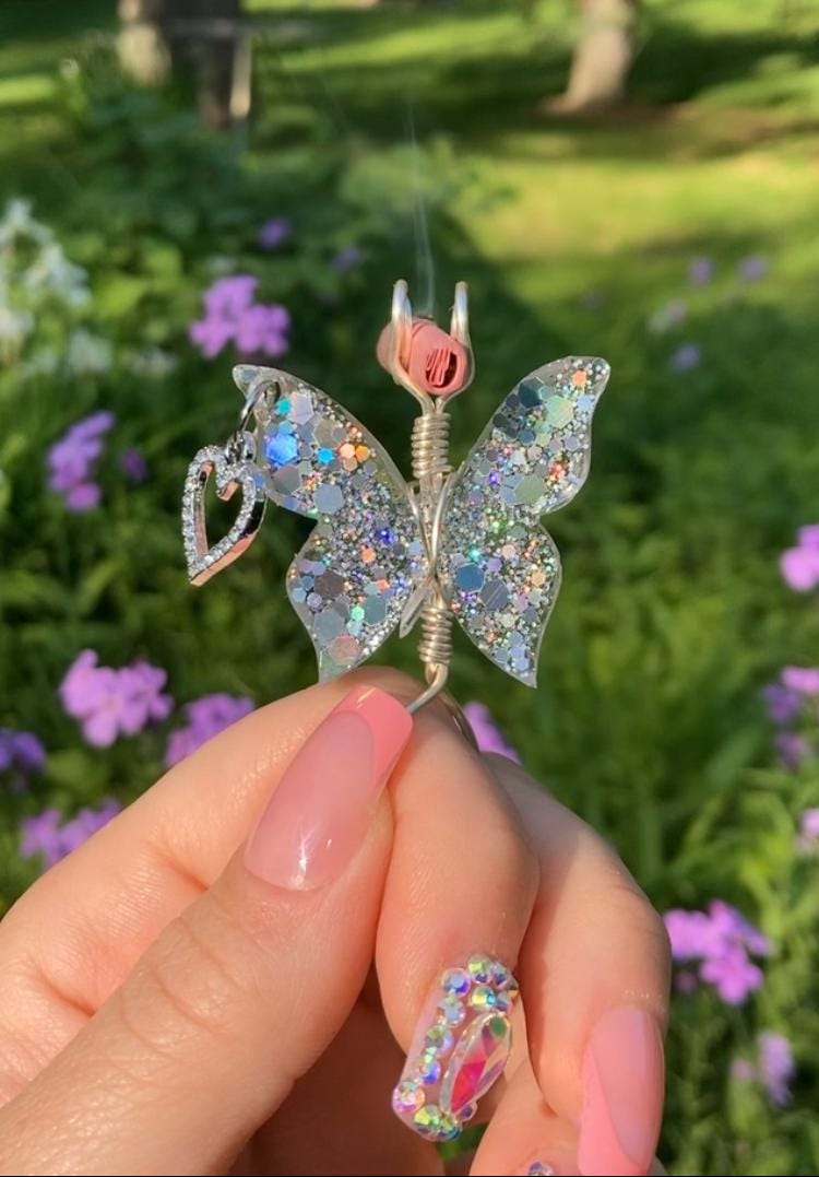 A hand with pink nails holds a sparkling, handmade butterfly joint holder featuring glittery wings, a heart charm.