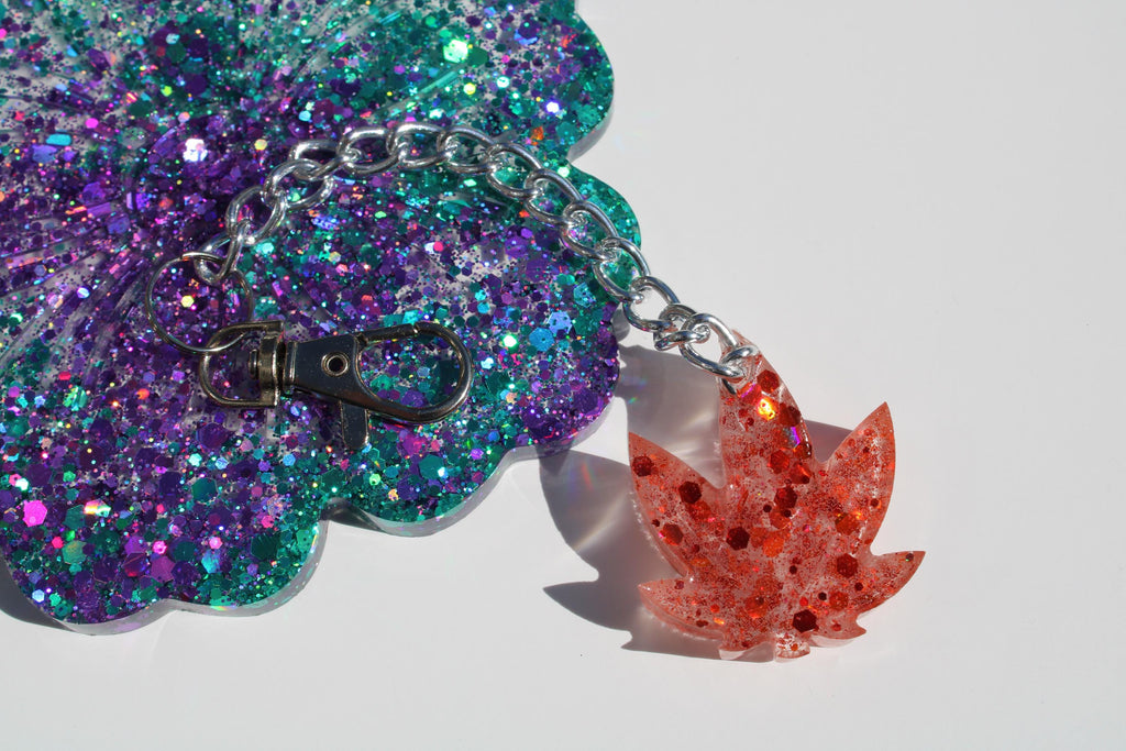 A sparkling purple and teal glittery resin coaster connects with a silver chain to a red marijuana leaf-shaped keychain, both handmade resin art accessories.