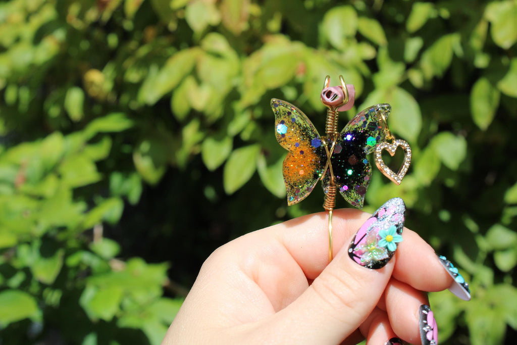 A hand with elaborate floral-themed nail art holds a decorative glittery butterfly smoke ring with a heart charm against a leafy green background.