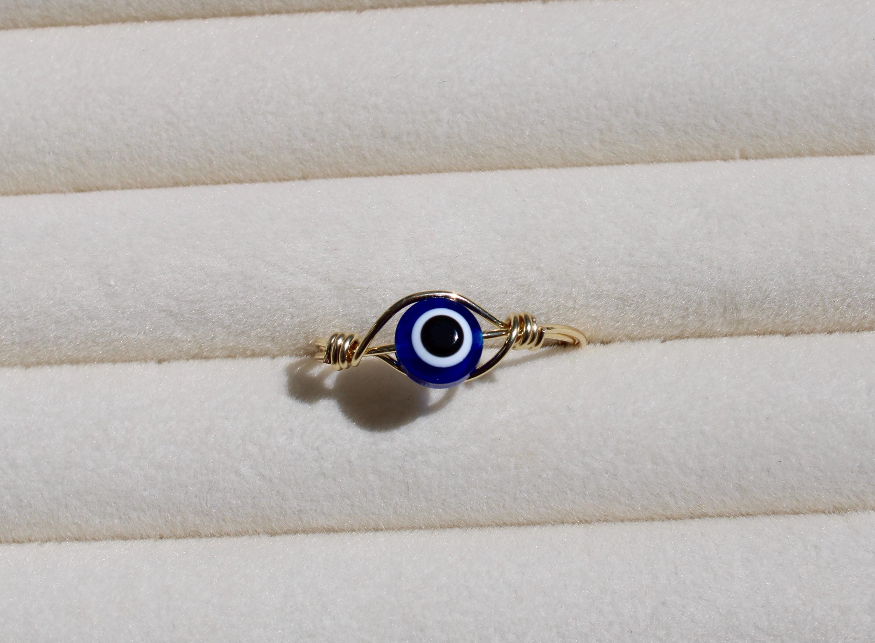 A close-up showcases a handcrafted gold wire ring featuring a blue and white Evil Eye bead against a soft, textured background.