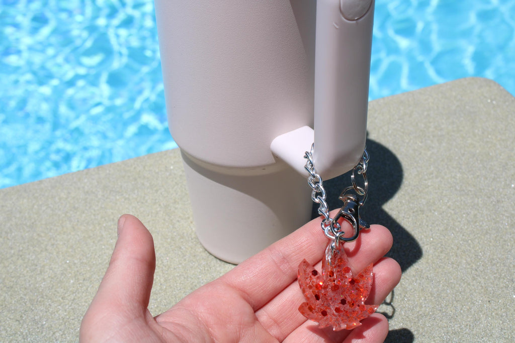A hand displays a sparkly red weed leaf keychain attached to a white tumbler against a poolside backdrop.