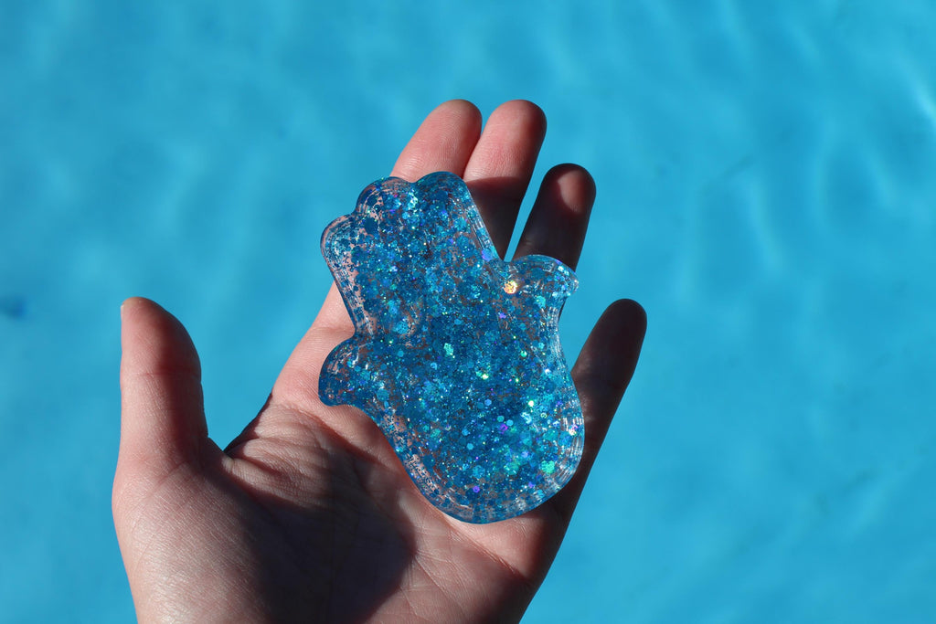 Hand holding a blue glittery Hamsa hand against a blue water background