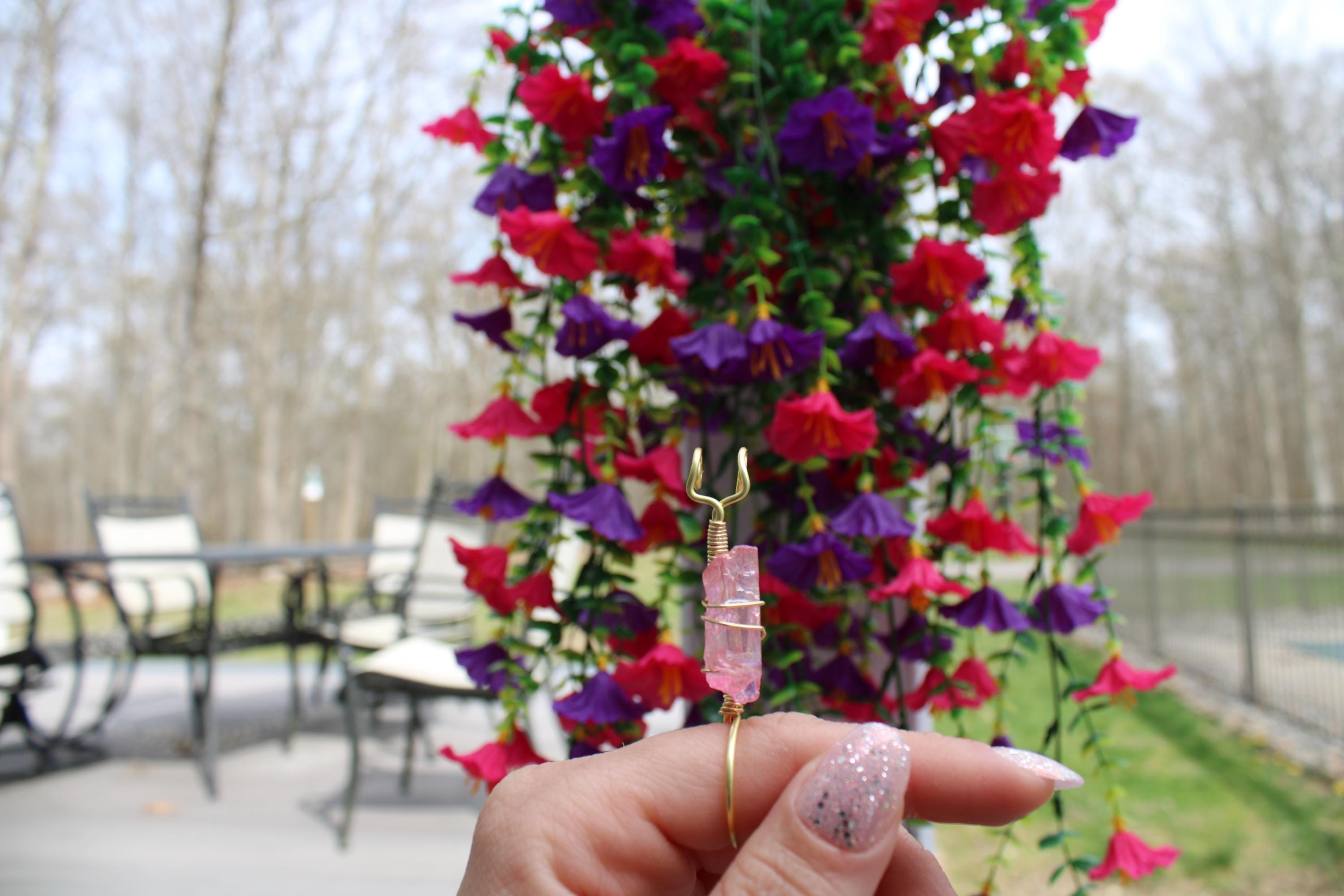 A hand with sparkly nails holds a raw pink crystal smoke ring with gold wire against a background of red and purple artificial flowers.