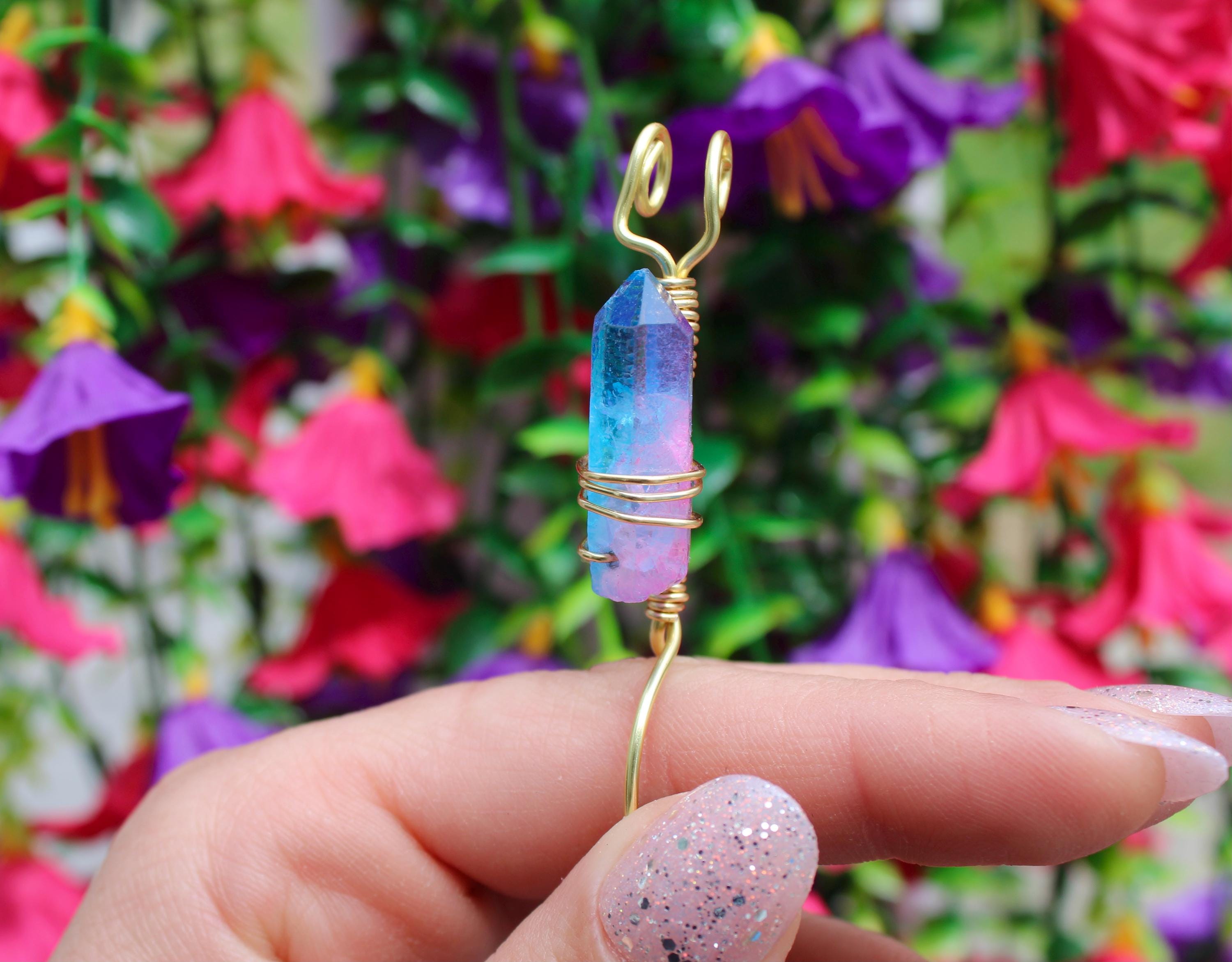 A manicured hand displays a blue and pink aura quartz crystal smoke ring wrapped in gold wire against a blurred floral background.