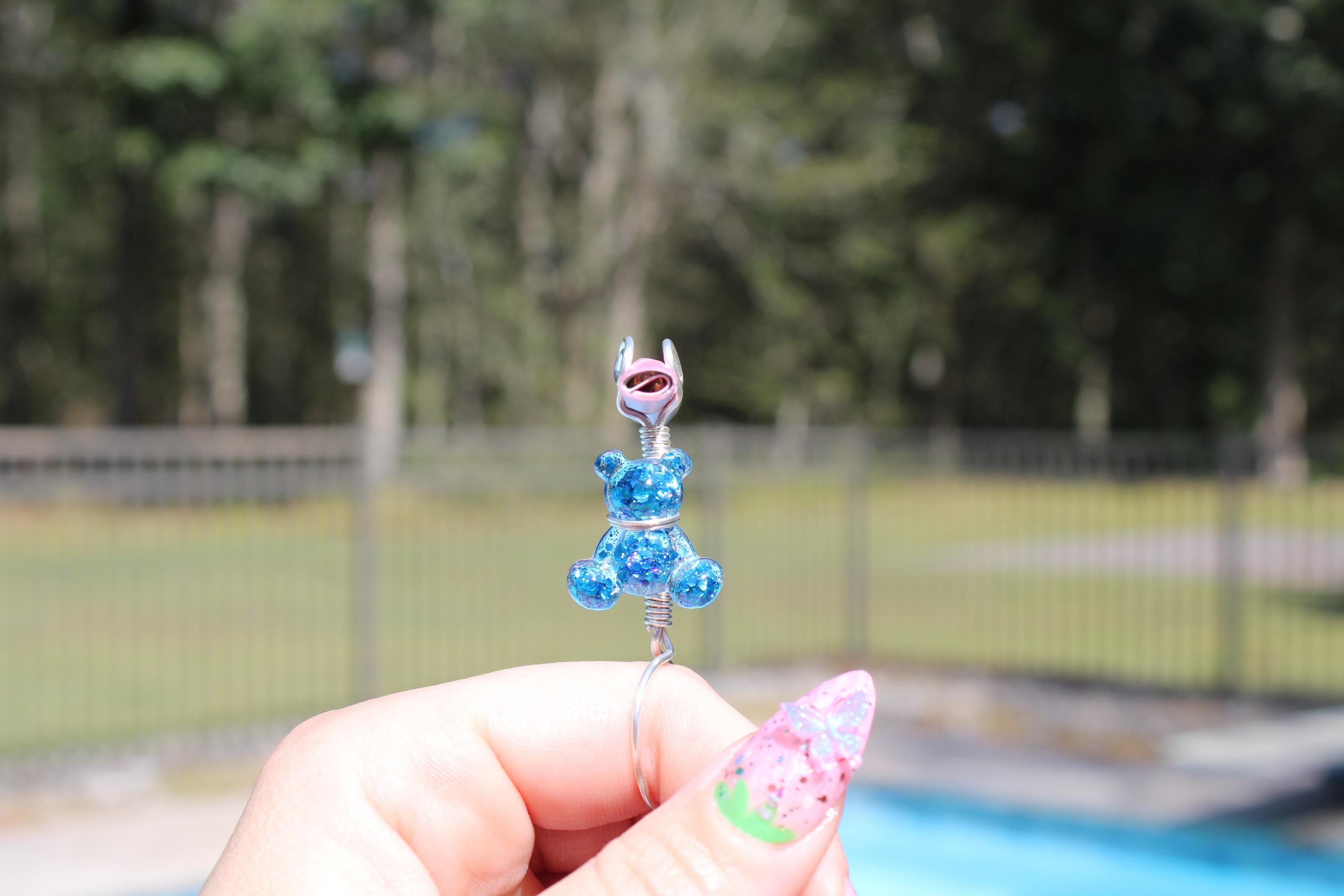 A hand with a decorated fingernail holds a handmade blue sequin teddy bear smoke ring in front of a pool.