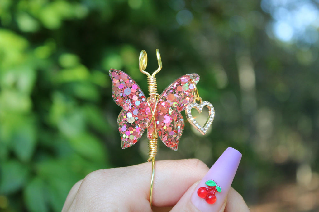 A hand displays a handcrafted, glittery pink butterfly smoke ring with a gold heart charm and wire wrap, complementing the lavender and cherry-decorated fingernails.