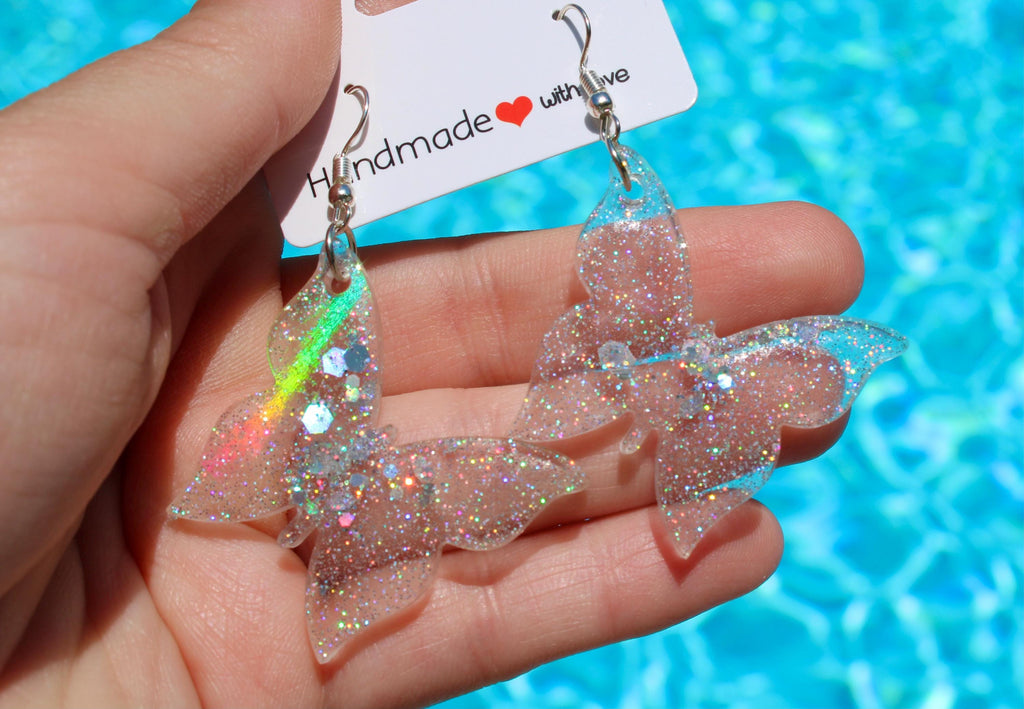 Someone showcases handmade butterfly earrings featuring shimmering glitter and iridescent accents against a bright blue pool backdrop.